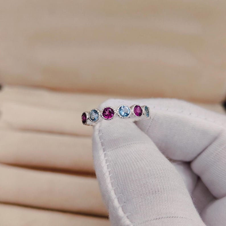 May include: A silver ring featuring a series of alternating gemstones. The ring showcases a pattern of round-cut gems in shades of pink, blue, and purple. The gemstones are set in a delicate, repeating design, creating a colorful and elegant piece of jewelry.