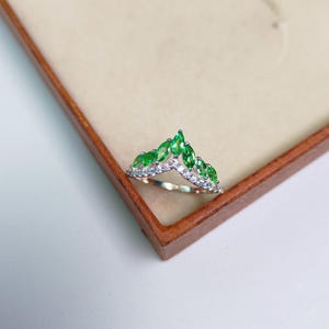 May include: A silver ring with a crown design, featuring marquise-cut green gemstones and small white stones. The ring is displayed in a wooden jewelry box, showcasing its elegant design and craftsmanship.