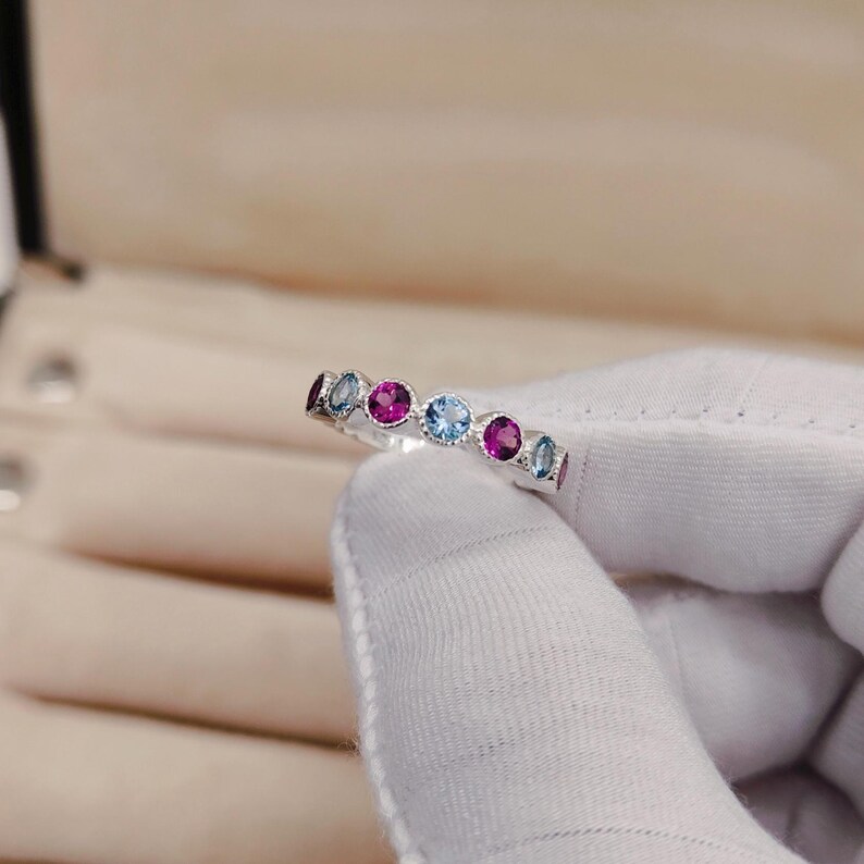 May include: A silver ring featuring a series of alternating round gemstones in shades of blue and magenta. The ring is held by a gloved hand, showcasing the colorful gemstones. The ring's design is simple, with each stone set closely together.