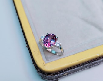 Può includere: Un anello d'argento con una grande pietra preziosa rosa scuro a taglio ovale come elemento centrale, affiancata da due pietre preziose rotonde e trasparenti più piccole. L'anello è incastonato in una fascia d'argento ed è esposto in un portagioie foderato di giallo.