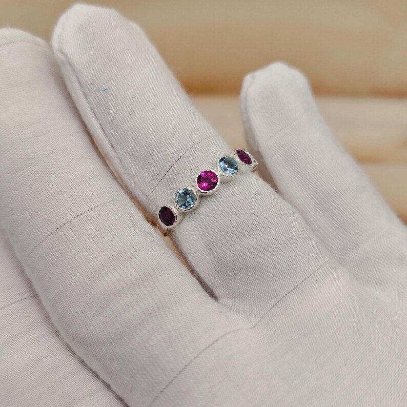 May include: A silver ring featuring a series of colorful gemstones. The ring showcases a row of round-cut gems in shades of red, blue, and pink. The gemstones are set in a polished silver band, creating a vibrant and eye-catching piece of jewelry.