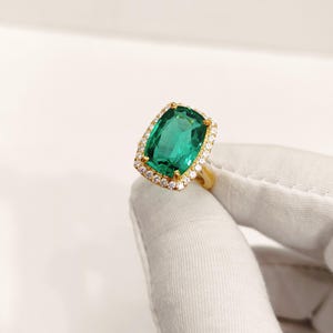 May include: A gold ring featuring a large, rectangular emerald-cut green gemstone. The gemstone is surrounded by a halo of smaller, clear gemstones. The ring is held by a gloved hand.