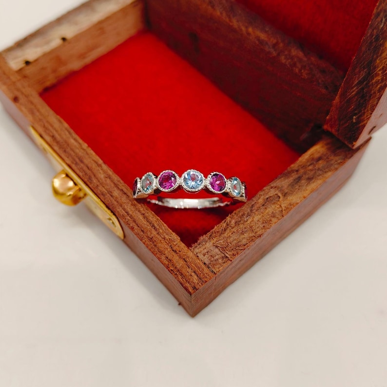 May include: A silver ring with a row of alternating pink and light blue gemstones. The ring is set in a wooden box with a red velvet interior. The gemstones are round and evenly spaced along the band.
