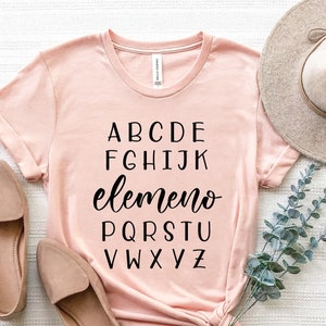 May include: A pink t-shirt with the alphabet printed on it. The letters are in black and the word "elemento" is written in cursive. The shirt is tied in a knot at the bottom.
