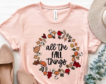 May include: A pink t-shirt with a graphic of a wreath of fall leaves, pumpkins, and acorns. The text "all the fall things" is inside the wreath.