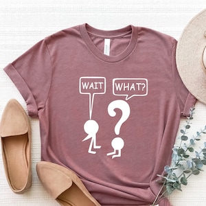 May include: A mauve t-shirt with a white graphic of two stick figures. The first stick figure is holding a speech bubble that says "WAIT" and the second stick figure is holding a speech bubble that says "WHAT?" with a question mark.