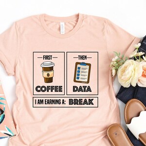 May include: A pink t-shirt with a graphic design that says "First Coffee, Then Data, I am earning a: Break". The graphic features a cup of coffee and a clipboard with a checklist.