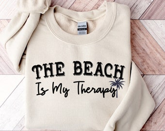 May include: A beige sweatshirt with the text "THE BEACH Is My Therapy" in black lettering and a palm tree graphic.