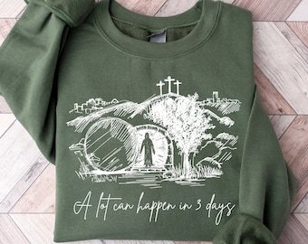 May include: A green sweatshirt with a white graphic of a tomb, a figure walking out of the tomb, and three crosses. The text "A lot can happen in 3 days" is printed below the graphic.