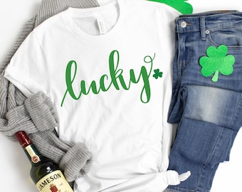May include: White t-shirt with the word "lucky" written in green cursive with a shamrock at the end. The shirt is laid out on a white surface with a gray sweater, blue jeans, and a bottle of Jameson Irish Whiskey.
