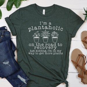 May include: A green t-shirt with the text "I'm a plantaholic on the road to recovery just kidding, I'm on my way to get more plants". The text is in white and there are three small drawings of potted plants.