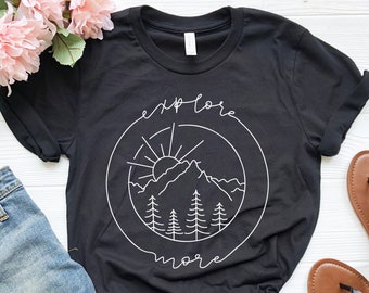 May include: Black t-shirt with a white graphic of a mountain range, sun, and trees inside a circle. The text "explore more" is written around the circle.