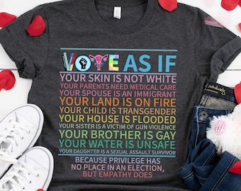 May include: A dark gray t-shirt with the text "Vote as if..." and a list of reasons to vote. The text is in rainbow colors and includes a fist, a uterus, and a voting symbol. The shirt is laid out on a white wooden surface with red rose petals scattered around it.
