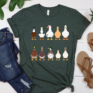 May include: A dark green t-shirt with a graphic of nine ducks in a row. The ducks are all facing away from the viewer and have different colored feathers. The t-shirt is on a white wooden surface.