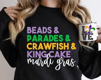 May include: A black sweatshirt with the text "BEADS & PARADES & CRAWFISH & KINGCAKE mardi gras" in green, yellow, and purple. A person is holding a white mug with a black fleur-de-lis design and the text "LOVE" in green, yellow, and purple.