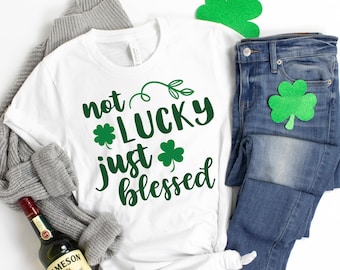May include: White t-shirt with green shamrock and text that reads "not lucky just blessed". The shirt is laid out on a white surface with a gray sweater, blue jeans, and green shamrock decorations.