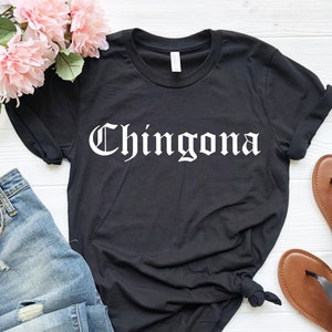 Op de afbeelding: Zwart T-shirt met het woord "Chingona" in wit gedrukt in een gotische lettertype. Het shirt wordt getoond op een wit oppervlak met blauwe jeans en bruine sandalen.