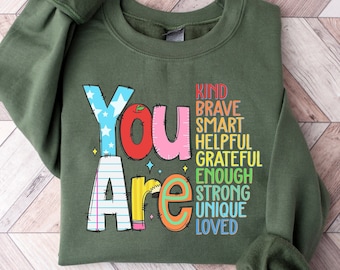May include: Green sweatshirt with the words "You Are" in colorful, stylized letters. The words "Kind, Brave, Smart, Helpful, Grateful, Enough, Strong, Unique, Loved" are stacked vertically in rainbow colors.