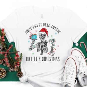 May include: White t-shirt with a skeleton wearing a Santa hat and holding a mug. The text on the shirt reads "When you're dead inside but it's Christmas".