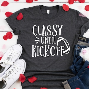 May include: A dark gray t-shirt with the text "Classy Until Kickoff" and a football graphic. The shirt is paired with blue jeans, white sneakers, and a stuffed animal. Red rose petals are scattered around the items.