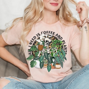 May include: A pink t-shirt with a graphic of a skeleton holding a cup of coffee surrounded by plants. The text on the shirt reads "All I need is coffee and plants".