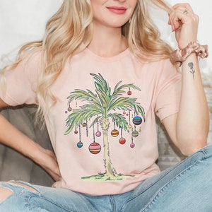 May include: A pink t-shirt with a graphic of a palm tree decorated with colorful ornaments. The palm tree is a whimsical take on a traditional Christmas tree.