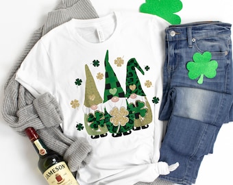 May include: A white t-shirt with a design of three gnomes wearing green hats and holding shamrocks. The gnomes are surrounded by green shamrocks and gold glitter.