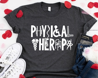 May include: A dark gray t-shirt with the text "PHYSICAL THERAPY" in white. The text is surrounded by white icons related to physical therapy, such as a dumbbell, a spine, a wheelchair, a walker, and a pair of crutches. The t-shirt is laid flat on a white surface with rose petals scattered around it.
