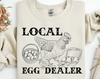 May include: A beige sweatshirt with a black graphic design of a chicken, eggs, and a basket. The text "LOCAL EGG DEALER" is printed in black below the graphic.