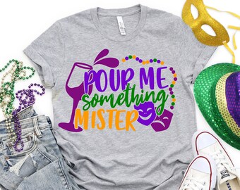 Puede incluir: Camiseta gris con el texto "Pour Me Something Mister" en morado, verde y naranja. El texto está rodeado de cuentas moradas y verdes y dos máscaras moradas y blancas.