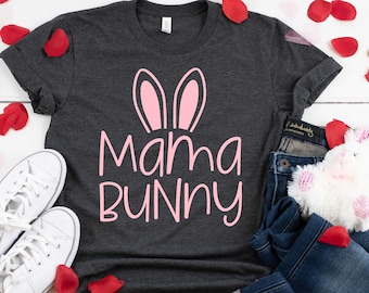 May include: A dark gray t-shirt with the words "Mama Bunny" in pink, featuring bunny ears. The shirt is paired with blue jeans, white sneakers, and a stuffed bunny. Red rose petals are scattered around the items.