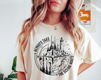 May include: A white t-shirt with a black graphic of a compass, mountains, a lighthouse, and waves. The text "Always Take The Scenic Route" is written around the compass.