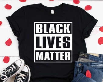 May include: Black t-shirt with the text "Black Lives Matter" in a white box on a black background. The shirt is laid out on a white wooden surface with red rose petals scattered around it. A pair of black canvas shoes and a pair of blue jeans are also in the image.