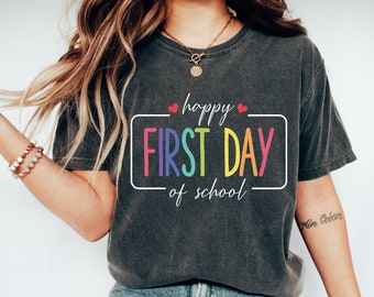 Puede incluir: Camiseta gris oscuro con el texto "Happy First Day of School" en una fuente colorida, similar al arcoíris. El texto está dentro de un borde rectangular blanco, con dos pequeños corazones rojos encima de la palabra "happy".