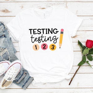 May include: White t-shirt with the text "TESTING testing 1 2 3" and a yellow pencil with a pink eraser.