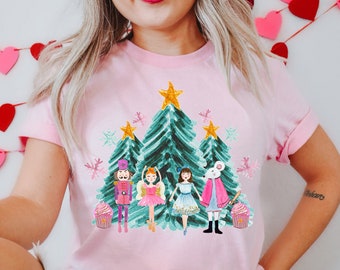 Könnte beinhalten: Ein pinkfarbenes T-Shirt mit einer Aquarell-Illustration eines Nussknackers, einer Ballerina, eines Mädchens in einem blauen Kleid und einer Maus in einem rosa Mantel, die vor drei Weihnachtsbäumen stehen. Die Bäume sind mit Schneeflocken und Sternen geschmückt.