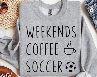 May include: A heather gray sweatshirt with the words "WEEKENDS COFFEE SOCCER" in black. A coffee cup and soccer ball graphic are included. The sweatshirt is displayed with brown shoes, sunglasses, and denim jeans.