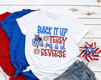 May include: White t-shirt with red, white, and blue text that reads "Back it up Terry put it in reverse". The text is accompanied by a graphic of a wheelchair with fireworks. The t-shirt is laid out on a white wooden surface with a pair of white sneakers, a blue t-shirt, a red t-shirt, and a pair of denim shorts.