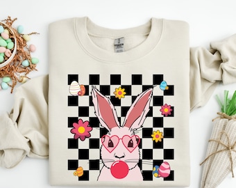 May include: A beige sweatshirt with a graphic of a pink bunny wearing heart-shaped sunglasses and blowing a bubble. The bunny is on a black and white checkered background with pink flowers and colorful Easter eggs.