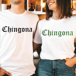 Op de afbeelding: Twee witte T-shirts met het woord "Chingona" erop gedrukt in zwarte en groene letters. De shirts worden gedragen door twee mensen.
