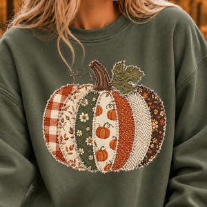 May include: An olive green crewneck sweatshirt featuring a large, central graphic of a pumpkin. The pumpkin is designed with a patchwork aesthetic, composed of various fabric patterns including plaid, floral, and herringbone in autumn colors.