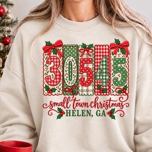 May include: A cream-colored crewneck sweatshirt featuring a graphic with the number 30545 in red and green patterns, decorated with bows and holly. Below the number, the text reads small town christmas HELEN, GA in red cursive and print.