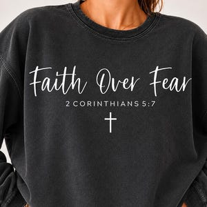 May include: A charcoal grey crewneck sweatshirt with a vintage-washed finish. The front features white text that reads Faith Over Fear, followed by 2 CORINTHIANS 5:7 and a small cross symbol centered below the scripture reference.