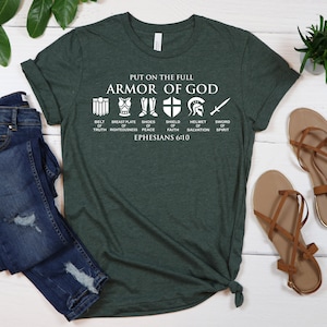 May include: A green t-shirt with a white graphic of the Armor of God. The graphic includes a belt, breastplate, shoes, shield, helmet, and sword. The text "PUT ON THE FULL ARMOR OF GOD" is above the graphic. The text "BELT OF TRUTH" is below the belt graphic. The text "BREASTPLATE OF RIGHTEOUSNESS" is below the breastplate graphic. The text "SHOES OF PEACE" is below the shoes graphic. The text "SHIELD OF FAITH" is below the shield graphic. The text "HELMET OF SALVATION" is below the helmet graphic. The text "SWORD OF SPIRIT" is below the sword graphic. The text "EPHESIANS 6:10" is below the graphic.