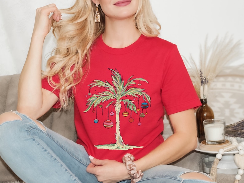 May include: Red t-shirt with a palm tree design and colorful ornaments hanging from the branches. The palm tree is a Christmas tree.