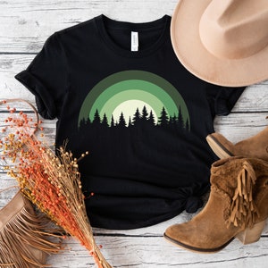 May include: Black t-shirt with a green and white forest silhouette design. The design features a stylized rainbow arch with trees in front of it.