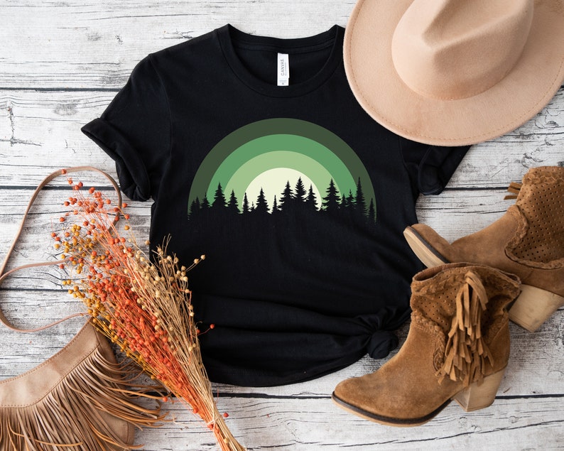 May include: Black t-shirt with a green and white forest silhouette design. The design features a stylized rainbow arch with trees in front of it.