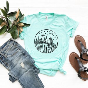 以下が含まれることがあります： ターコイズブルーのTシャツに、月と星が描かれた円の中にホグワーツ城の白黒のグラフィックがプリントされています。グラフィックはTシャツの中央に配置されています。