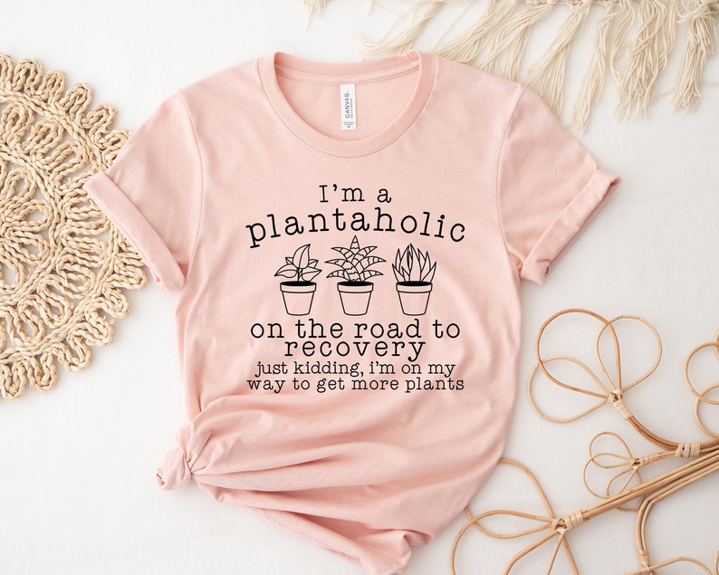May include: A pink t-shirt with a black graphic that says "I'm a plantaholic on the road to recovery just kidding, I'm on my way to get more plants". The graphic includes three potted plants.