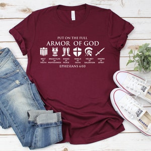 May include: A maroon t-shirt with white text that reads "Put on the full Armor of God" and includes a graphic of the armor of God with the text "Belt of Truth, Breastplate of Righteousness, Shoes of Peace, Shield of Faith, Helmet of Salvation, Sword of Spirit, Ephesians 6:10".